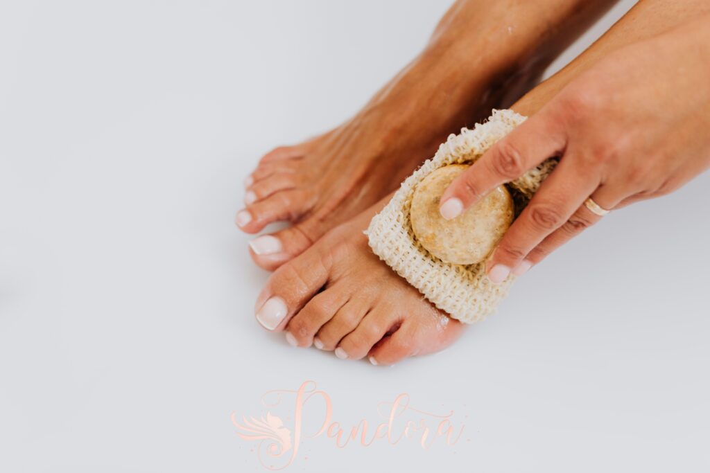 Close-up of a woman exfoliating her feet with a pumice stone and scrub pad, emphasizing personal car — Pandora Nail Spa, Lafayette, CA